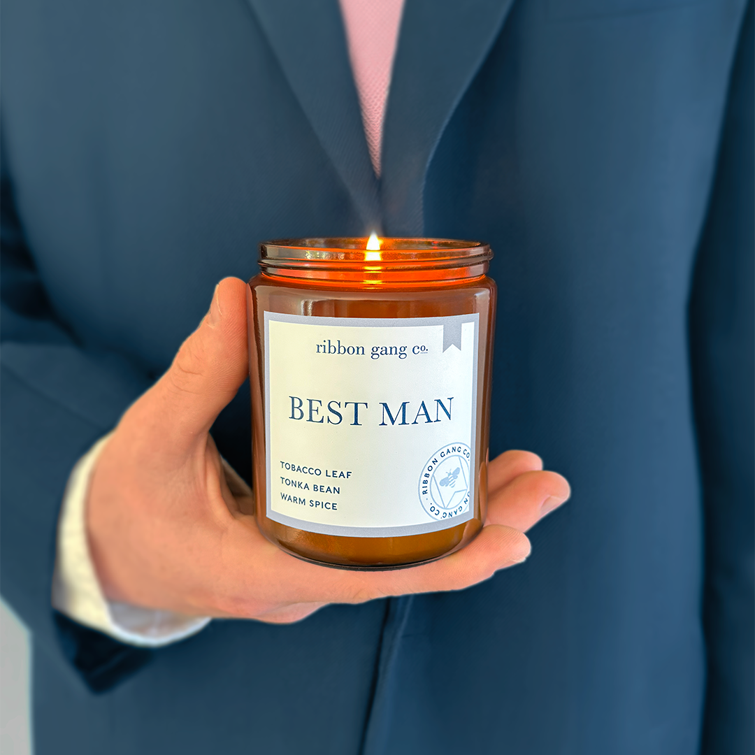 A man in a blue suit with a pale pink tie holding a lit Best Man candle in an amber jar.