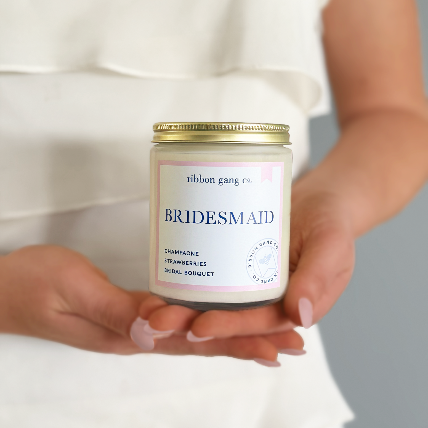 Candle jar labeled 'Bridesmaid' held by hands against a neutral background.