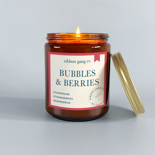 Candle in a glass jar with a label reading 'Bubbles & Berries' by Ribbon Gang Co. on a grey background.