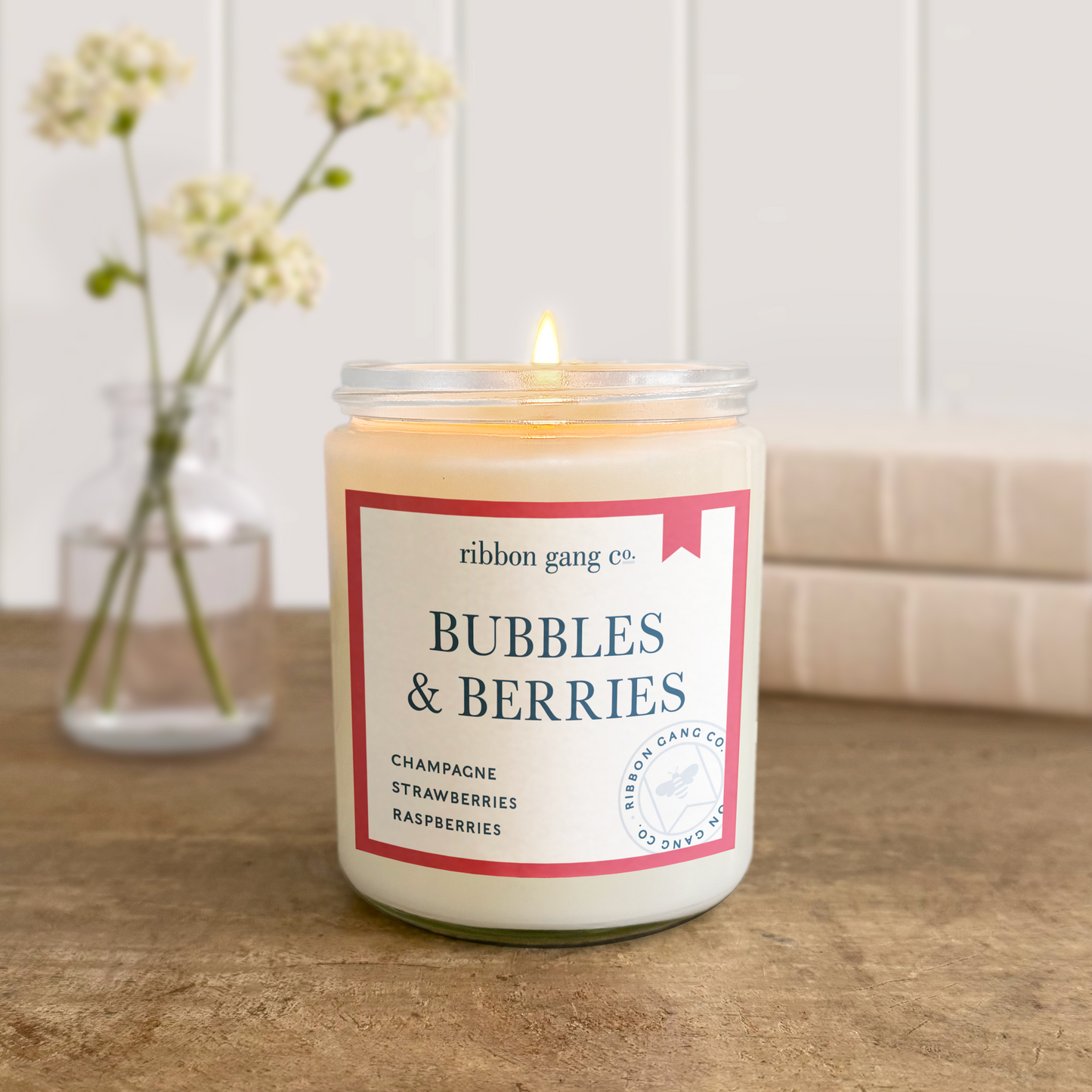Candle labeled 'Bubbles & Berries' by Ribbon Gang Co. on a wooden surface with a vase of flowers in the background.