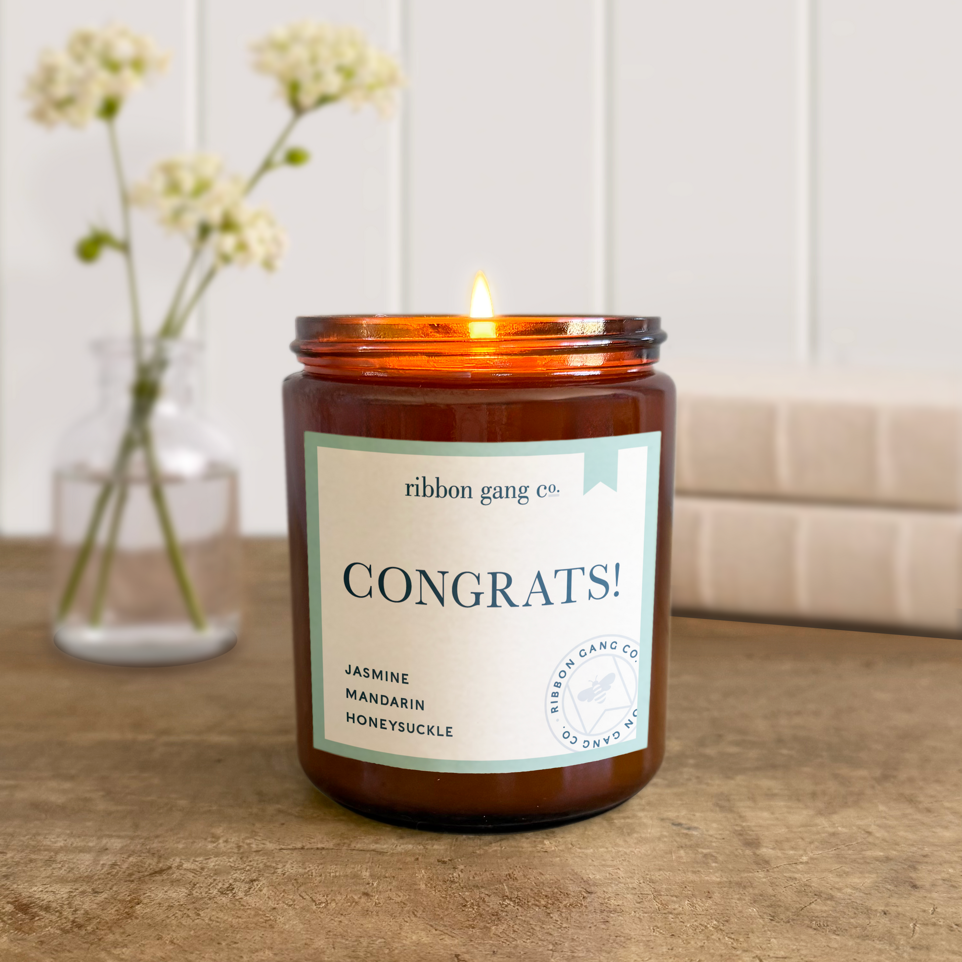 Candle with 'Congrats!' label on a wooden surface, next to a vase with flowers.