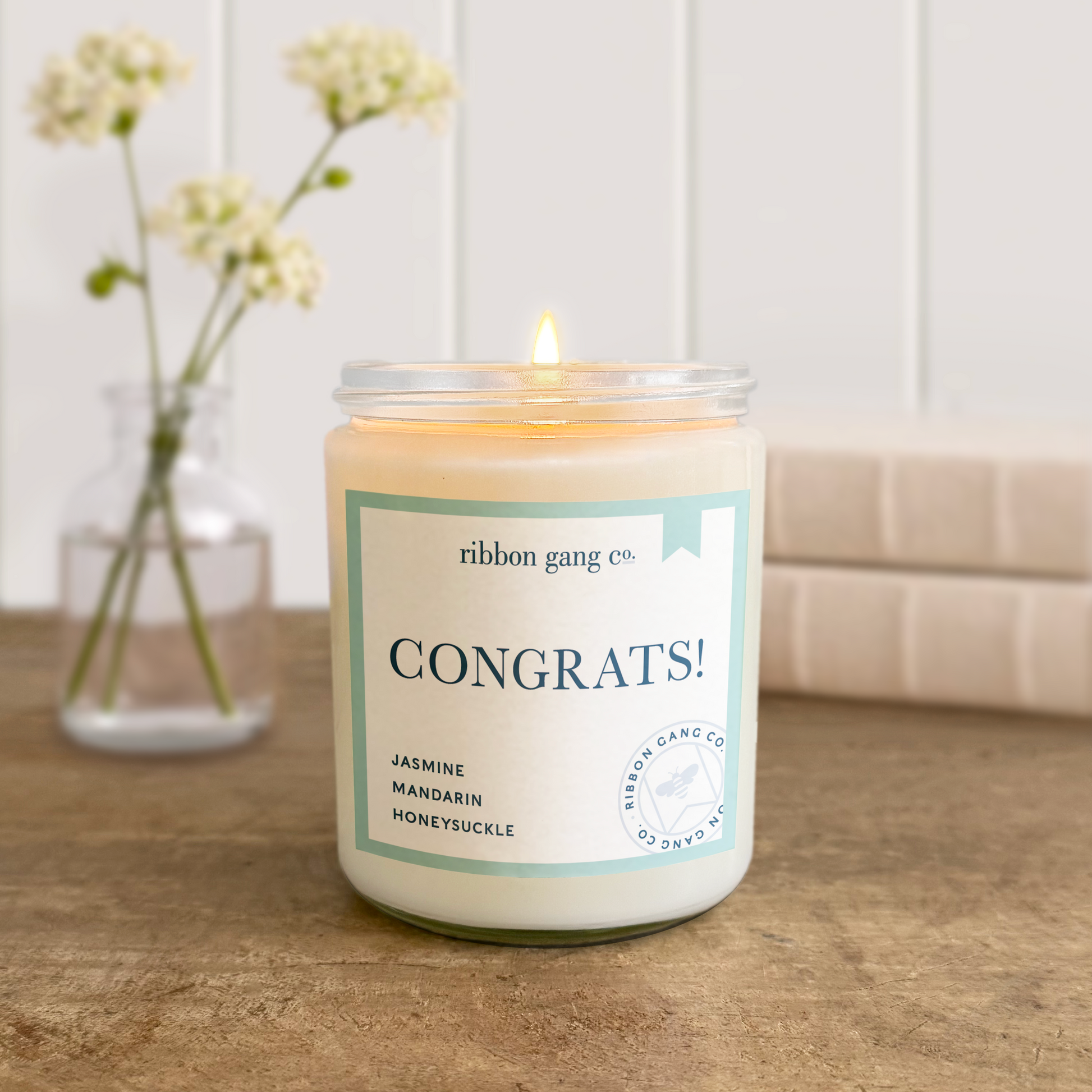 Candle with 'Congrats!' label on a wooden surface, next to a vase with flowers.