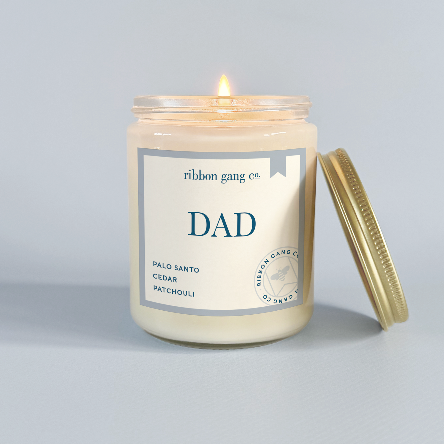 A Dad candle in a clear jar lit with a gold lid resting against it on a pale blue background.