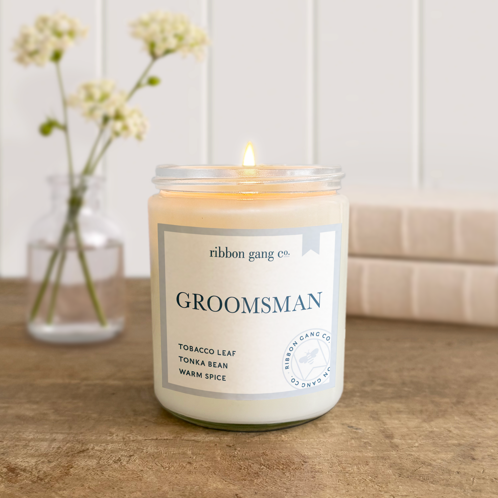 Candle with 'Groomsman!' label on a wooden surface, next to a vase with flowers.