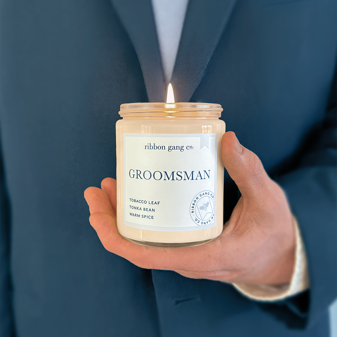 A man in a blue suit with a pale blue tie holding a lit Groomsman candle in a clear jar.
