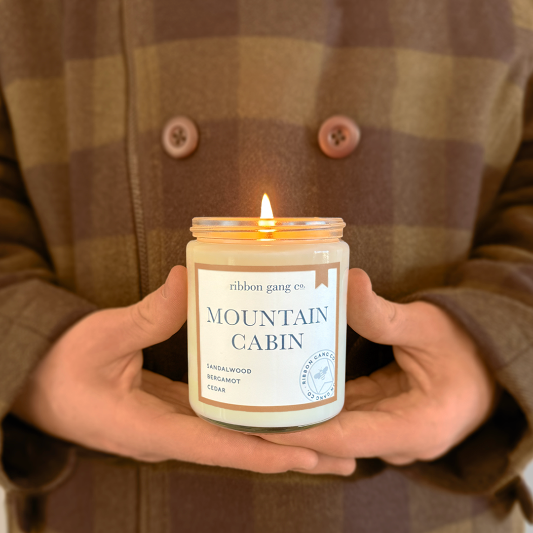 A person in a brown plaid coat holds a lit candle in a clear glass jar. The candle label reads 'Mountain Cabin'.