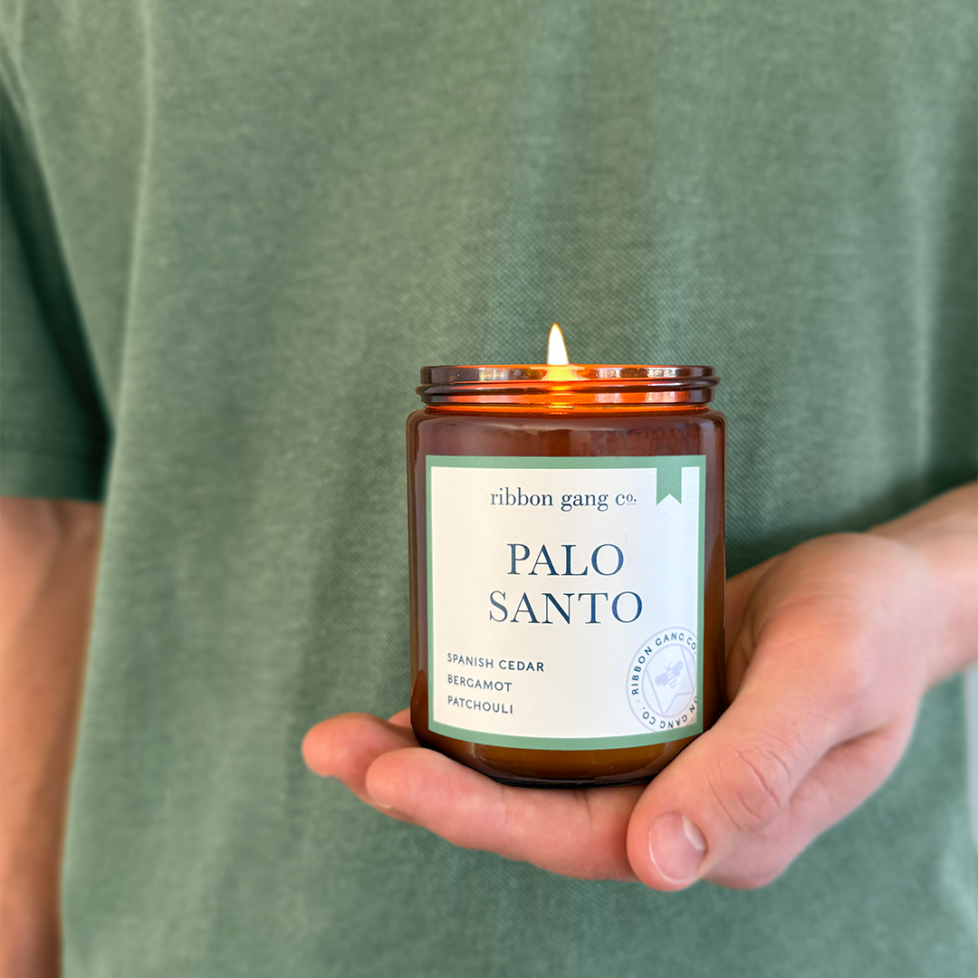 A person in a green shirt holds a lit amber glass candle jar labeled 'Palo Santo' by Ribbon Gang Co. against a plain light background.