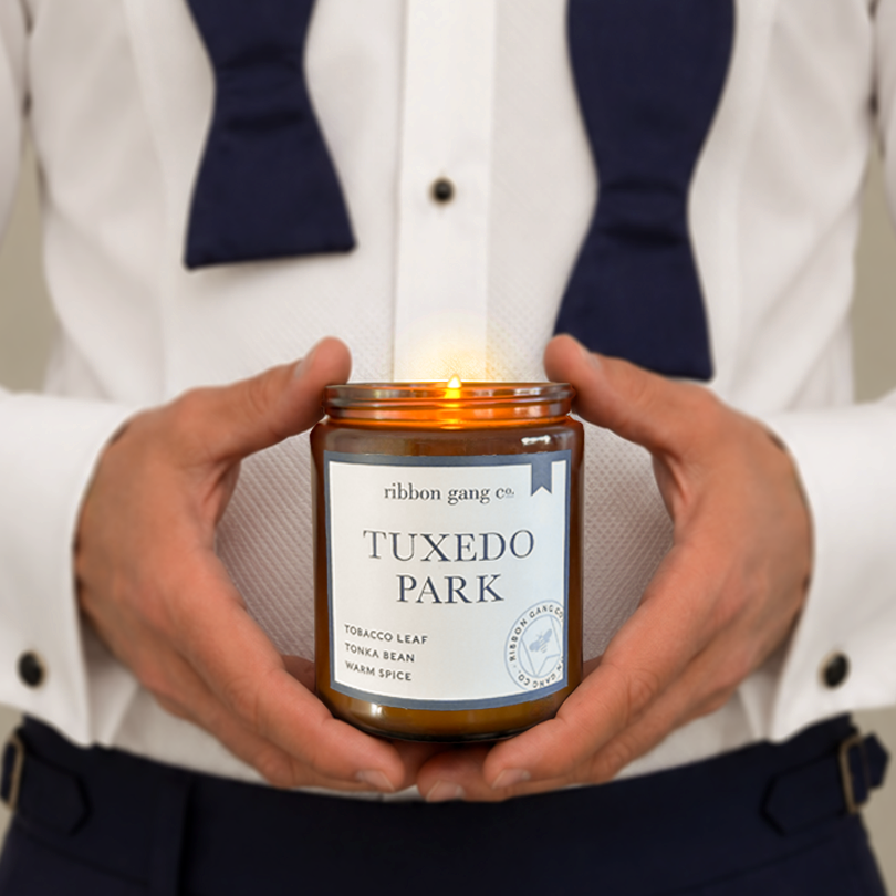 A lit amber glass jar candle held in both hands by a person wearing a white dress shirt, navy bow tie, and navy trousers. 