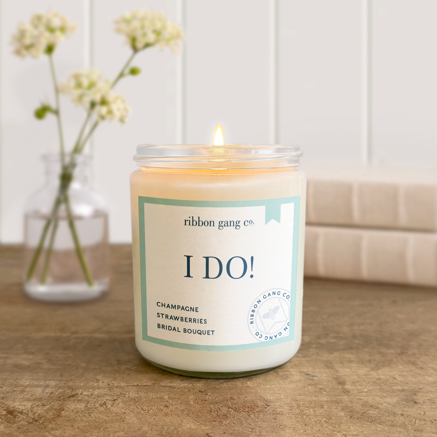 Candle with 'I DO!' label on a wooden surface, next to a vase with flowers.