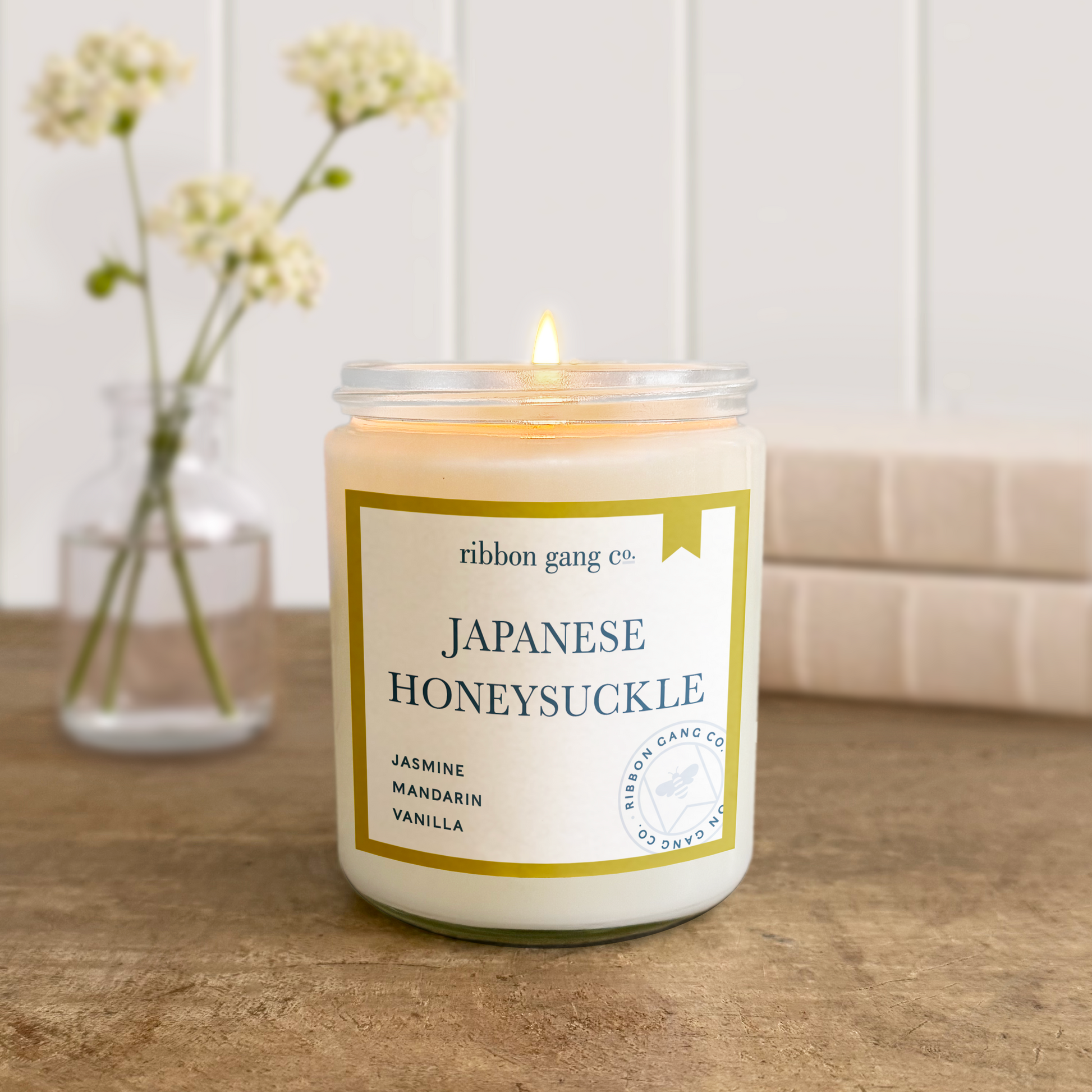 Candle labeled 'Japanese Honeysuckle' by Ribbon Gang Co. on a wooden surface with a vase of flowers in the background.