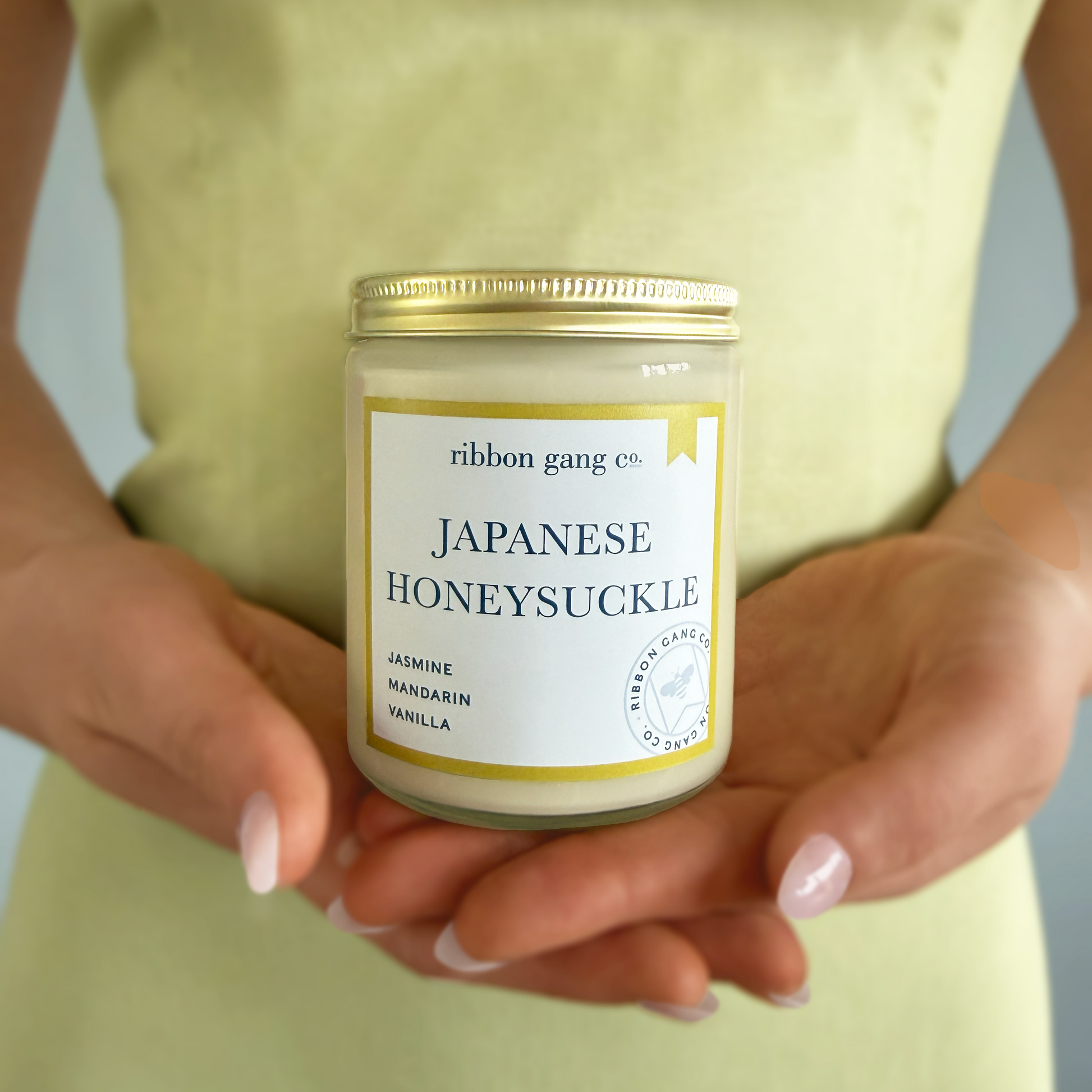 Person holding a candle labeled 'Japanese Honeysuckle' by Ribbon Gang Co.