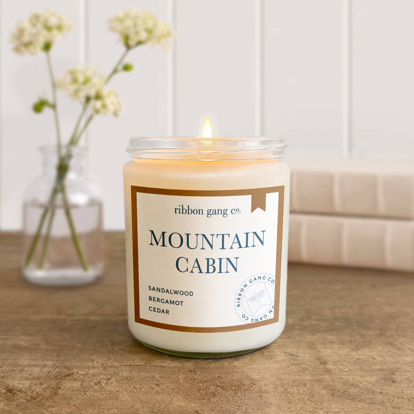 Candle labeled 'Mountain Cabin' by Ribbon Gang Co. on a wooden surface with a vase of flowers in the background.