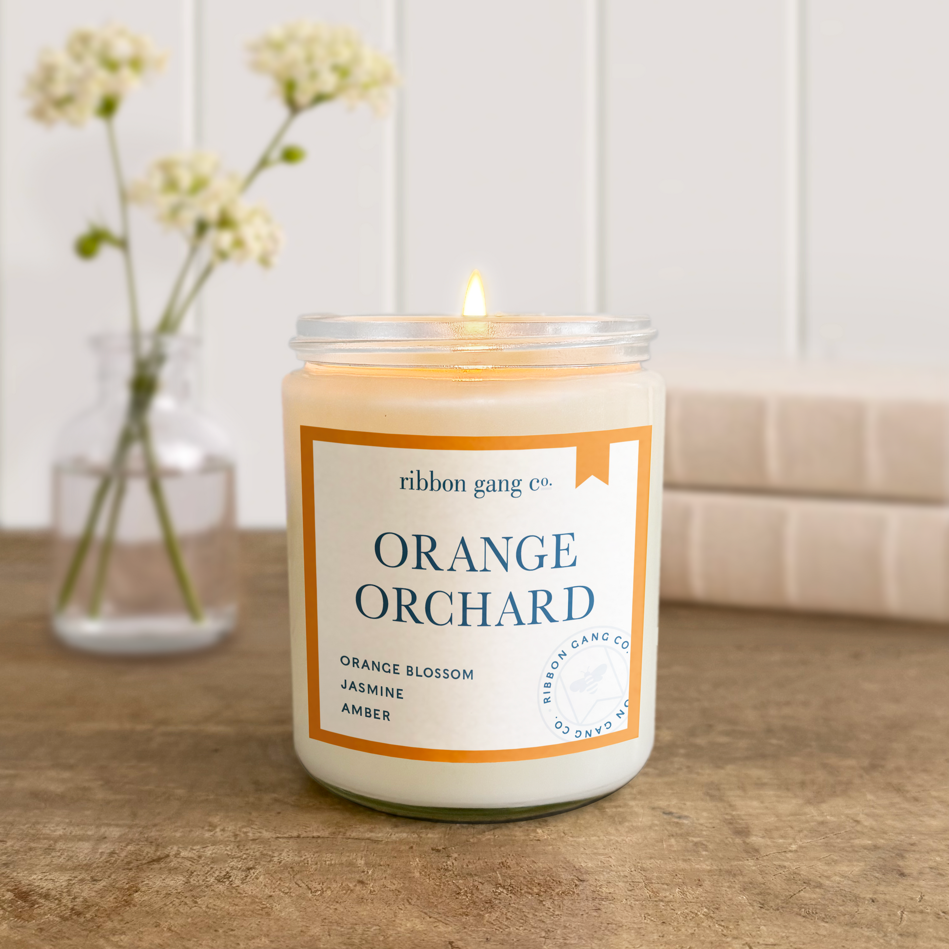 Candle labeled 'Orange Orchard' by Ribbon Gang Co. on a wooden surface with a blurred background.