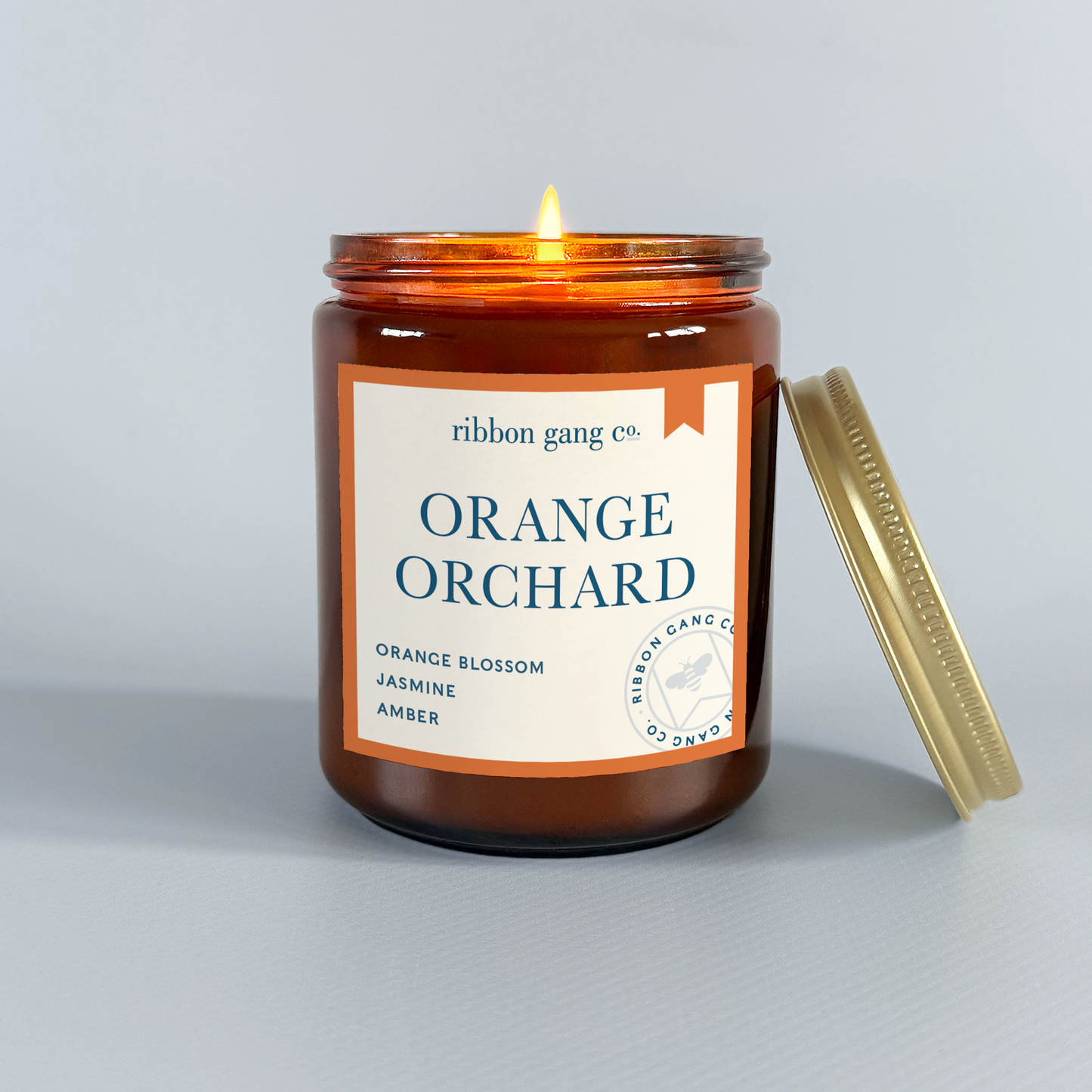 Candle in an amber jar labeled 'Orange Orchard' by Ribbon Gang Co. on a light grey background.