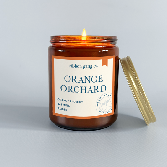 Candle in an amber jar labeled 'Orange Orchard' by Ribbon Gang Co. on a light grey background.