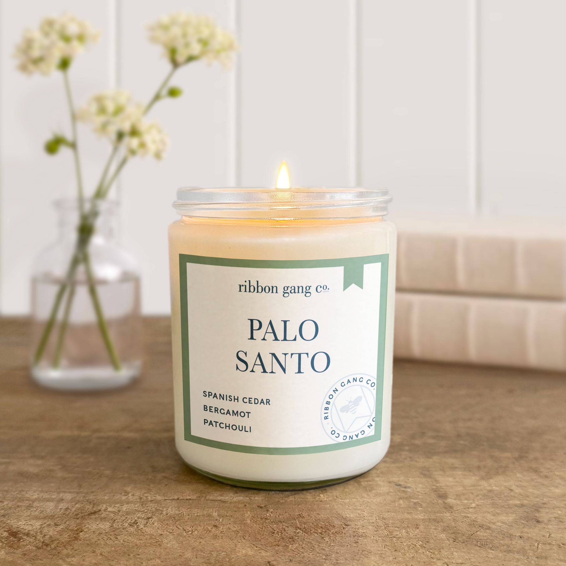 Candle labeled 'Palo Santo' by Ribbon Gang Co. on a wooden surface with a vase of flowers in the background.