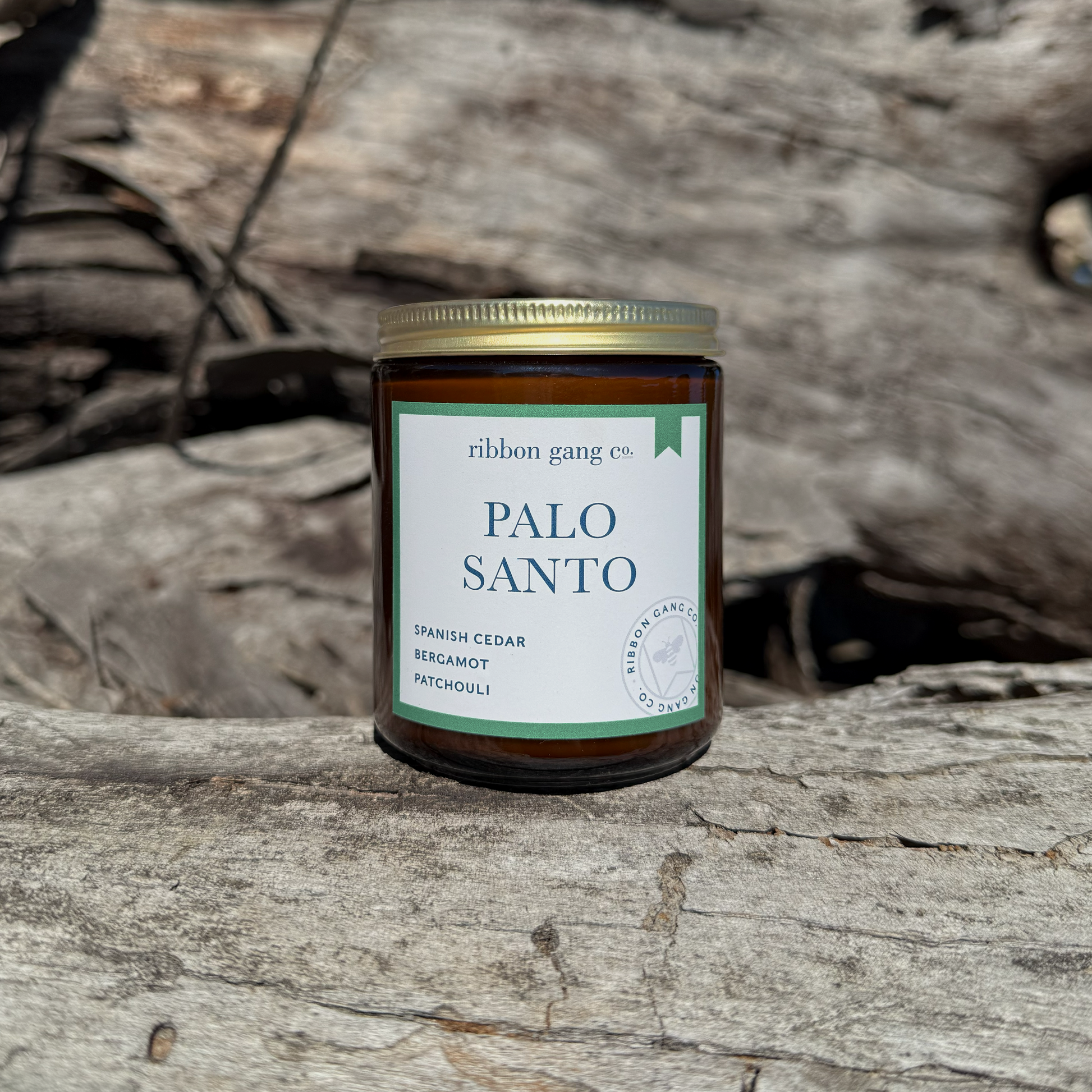 Candle jar labeled 'Palo Santo' by Ribbon Gang Co. on a woden log with wooden logs behind it.