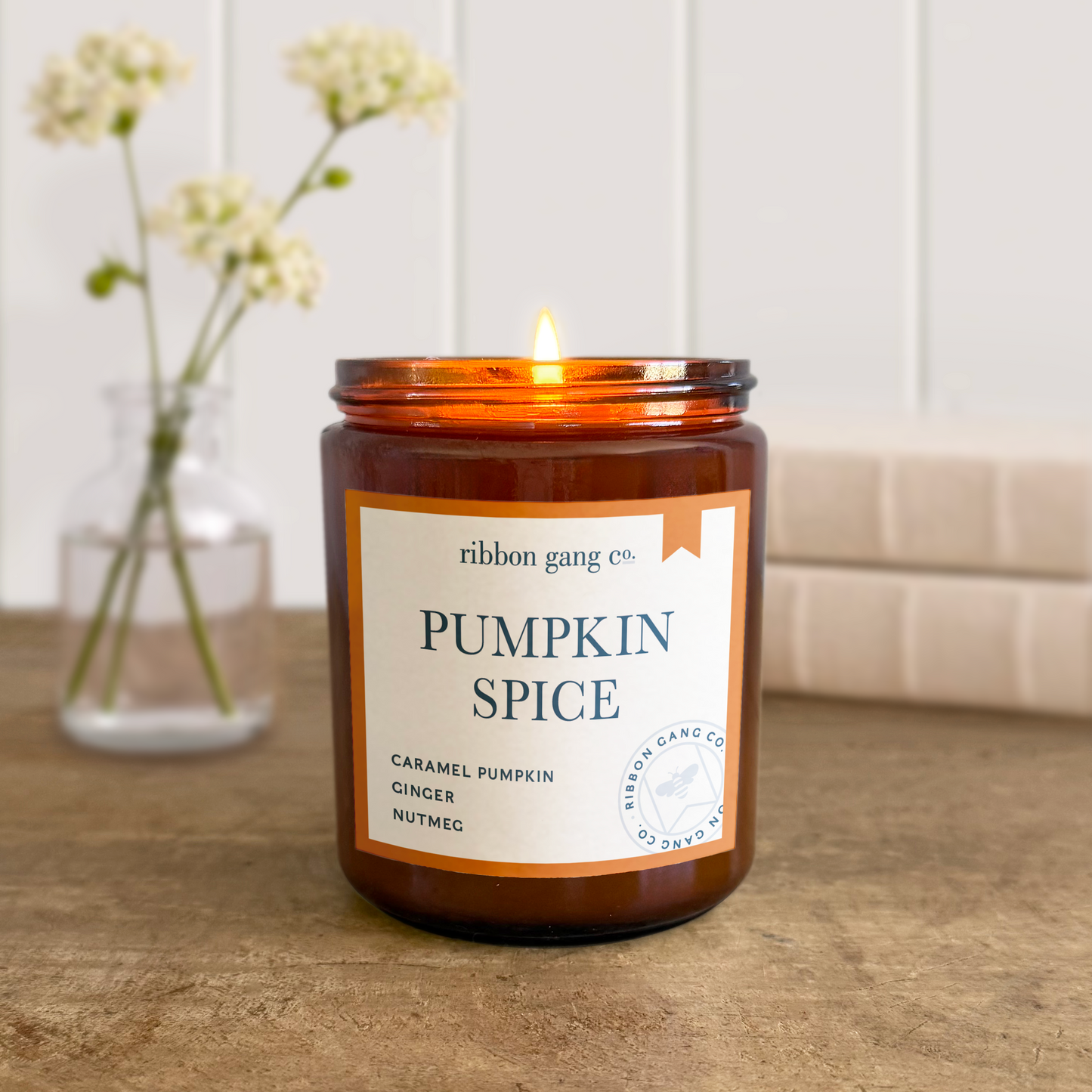 Pumpkin Spice candle burning in an amber jar on a wooden table with flowers in the background.