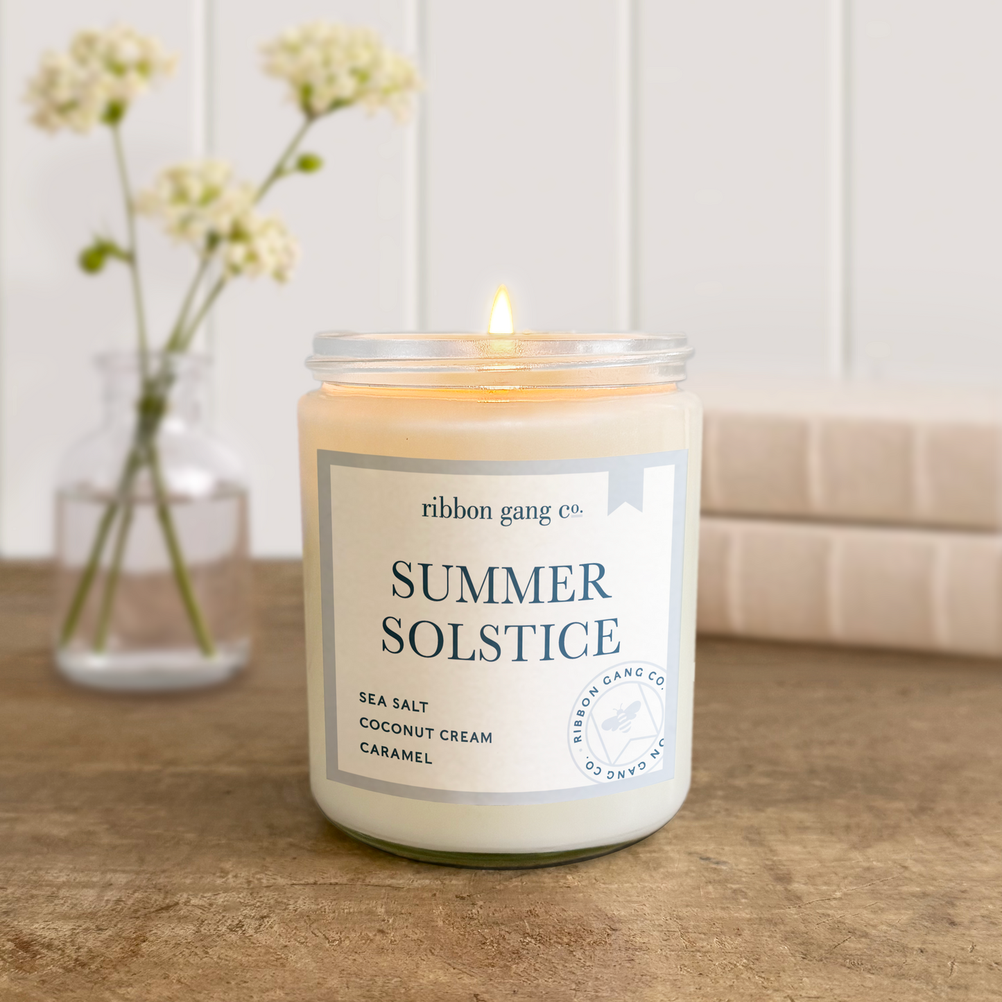 Candle labeled 'Summer Solstice' by Ribbon Gang Co. on a wooden surface with a vase of flowers in the background.