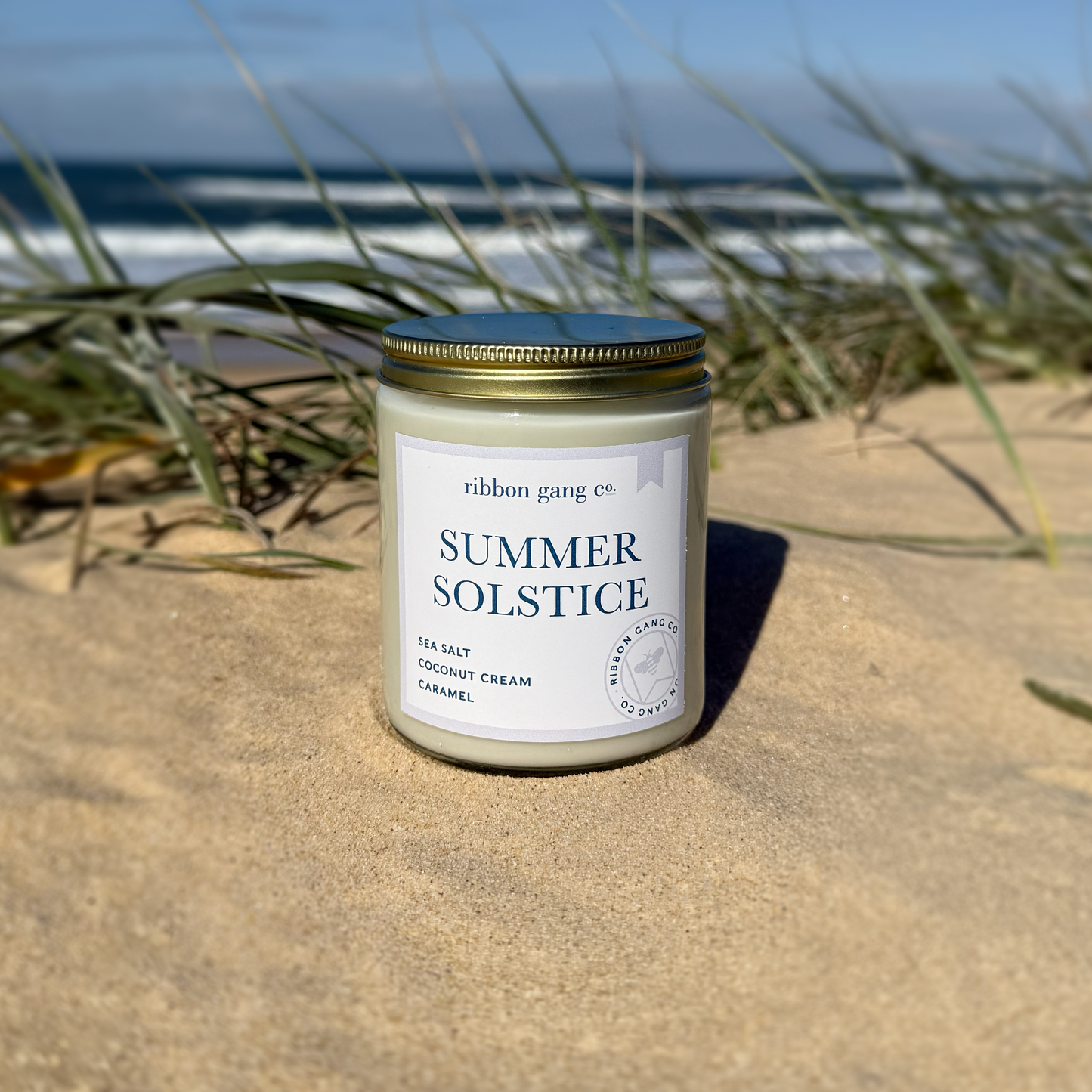 Candle labeled 'Summer Solstice' on beach with grass and water in the background.