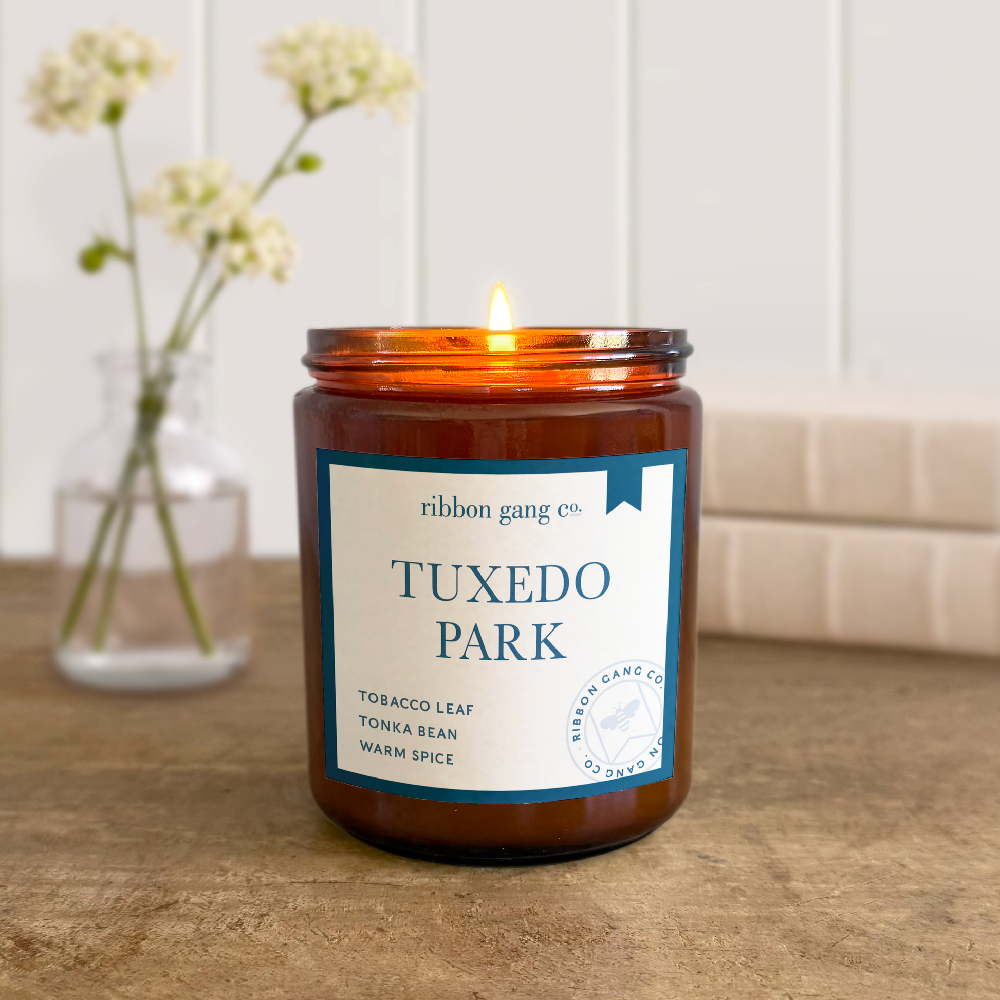 Candle with a label reading 'Tuxedo Park' on a wooden surface, with a vase of flowers in the background.