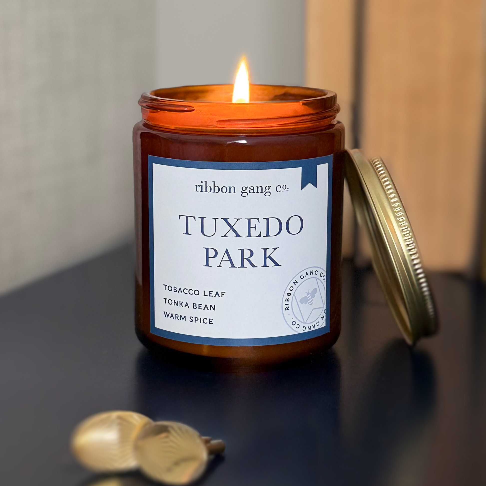 Candle in a jar labeled 'Tuxedo Park' by Ribbon Gang Co. on a navy surface with two gold cufflinks.