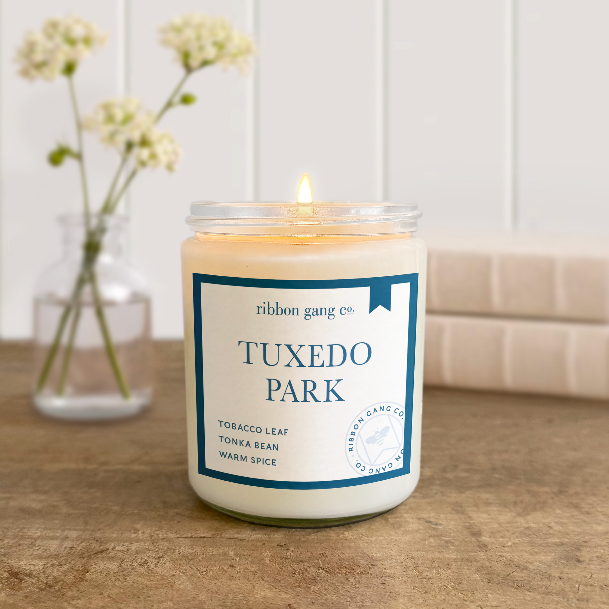 Candle with a label reading 'Tuxedo Park' on a wooden surface, with a vase of flowers in the background.
