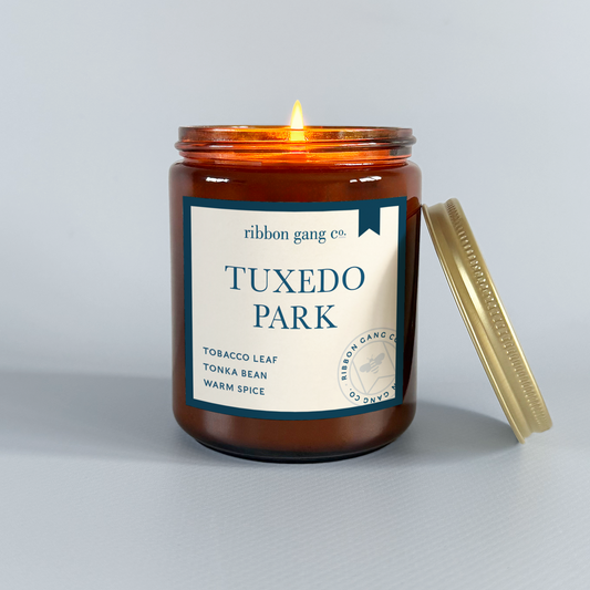 Candle in a jar labeled 'Tuxedo Park' by Ribbon Gang Co. on a grey background.