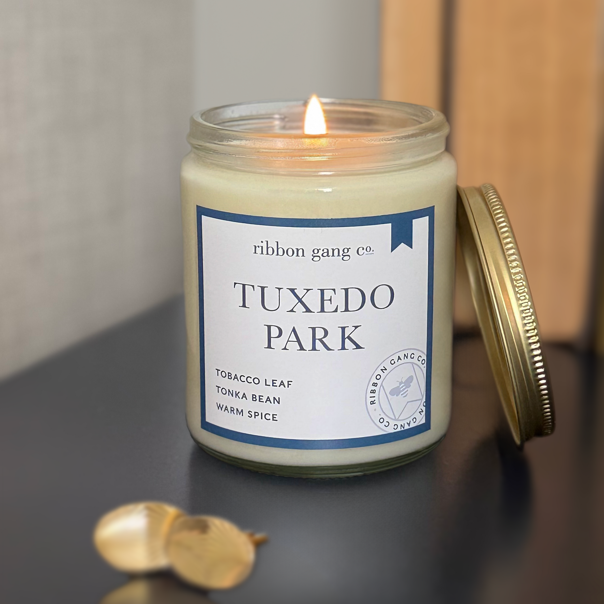 Candle in a jar labeled 'Tuxedo Park' by Ribbon Gang Co. on a navy surface with two gold cufflinks.